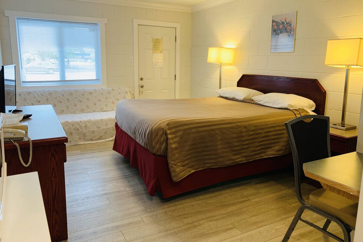 A hotel room with a bed, bedside lamps, a desk with a telephone, a chair, a sofa, and a window with blinds.