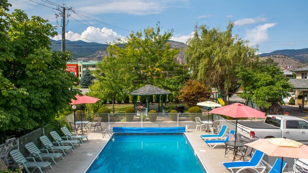 A swimming pool with lounge chairs and umbrellas, surrounded by greenery and a view of mountains. - Travelers Motel (Penticton)