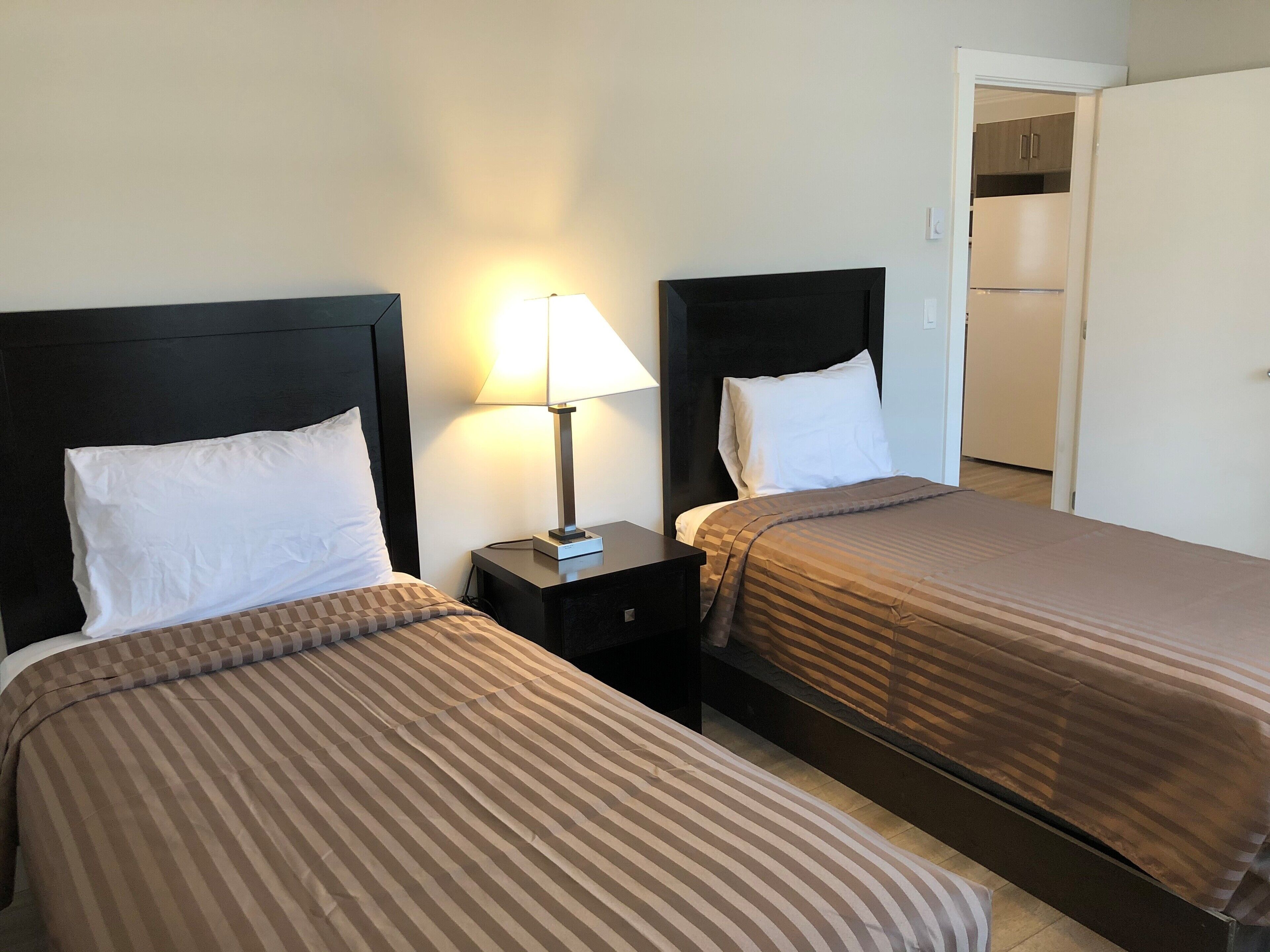 Quarto de hotel com duas camas, cada uma com colcha listrada e travesseiros brancos. Há uma mesinha de cabeceira com abajur entre as camas. Uma área de cozinha é visível ao fundo.
