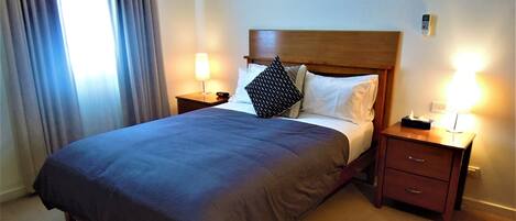 A neatly made bed with a dark blue bedspread, two pillows, and a wooden headboard. There are two bedside tables with lamps, one on each side of the bed.