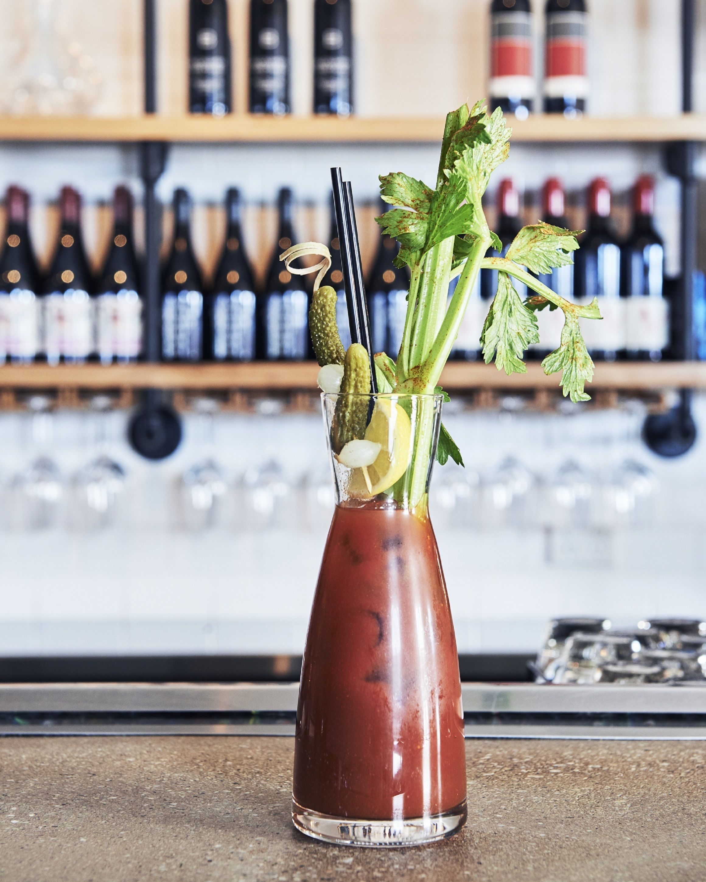 A Bloody Mary cocktail in a glass with celery, lemon, and olives, with a blurred background of wine bottles.