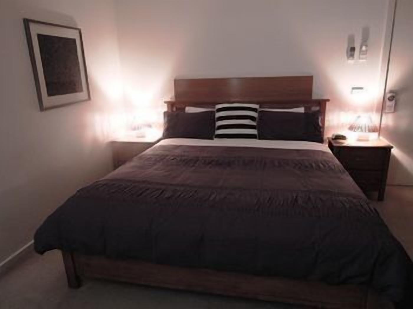 A bedroom with a wooden bed, two bedside tables, a framed picture on the wall, and a bedside lamp.