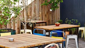 Ein Außenbereich mit einer Terrasse, Holztafeln, Stühlen, einem kleinen Topfpflanzen und einem Holzzaun im Hintergrund.