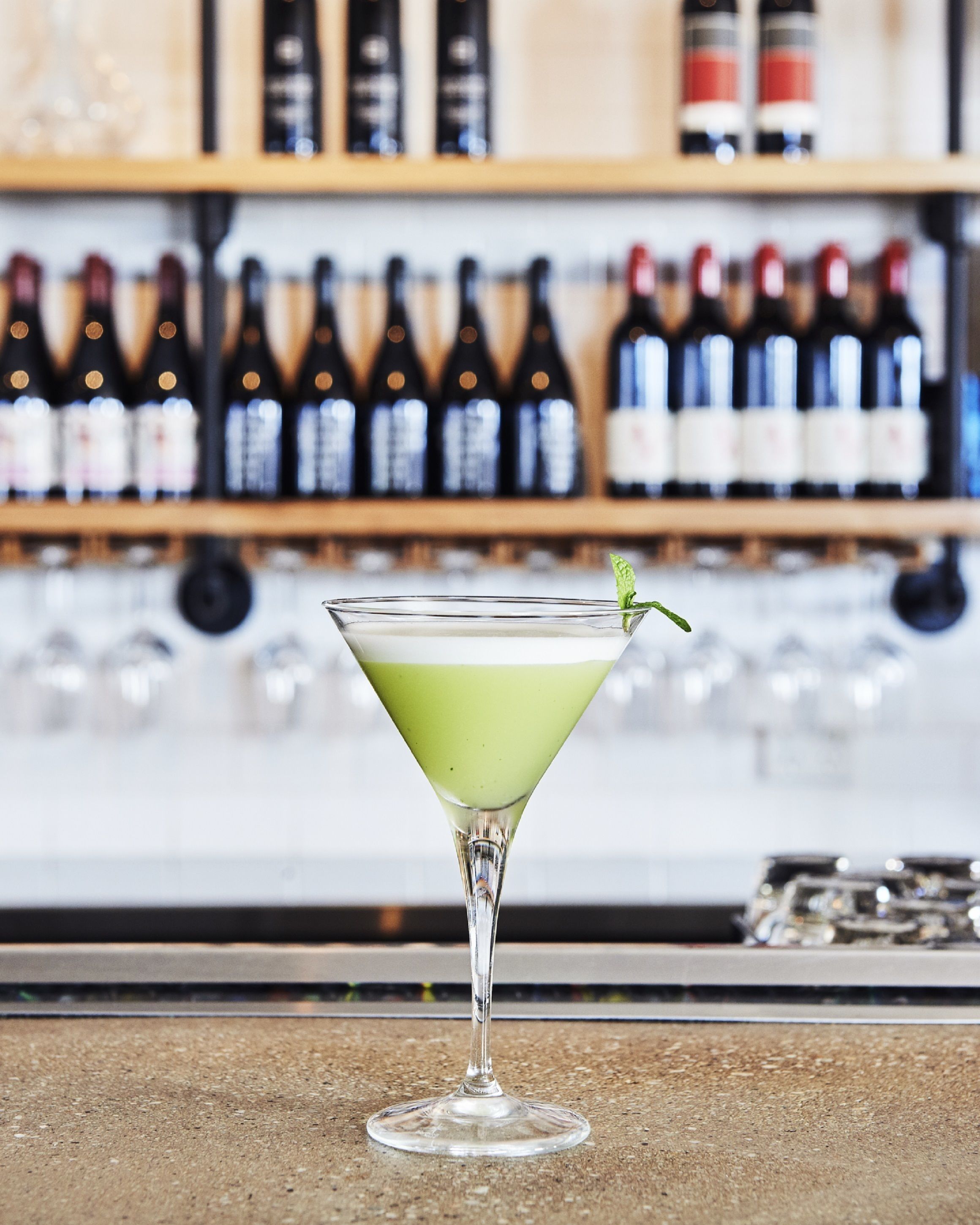 Un verre à martini, décoré d’une feuille de menthe, posé sur le comptoir d’un bar, avec un arrière-plan flou compos é de bouteilles de vin.