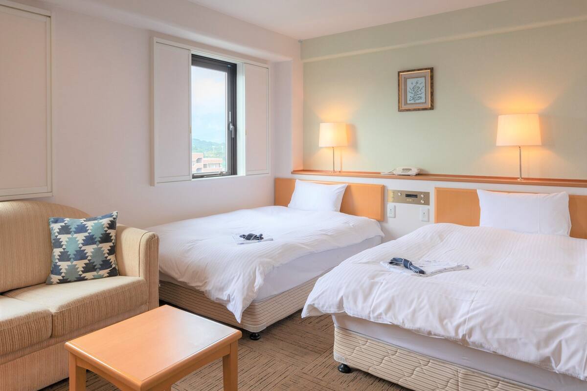 A hotel room with two beds, a sofa, a small table, and a window with a view.