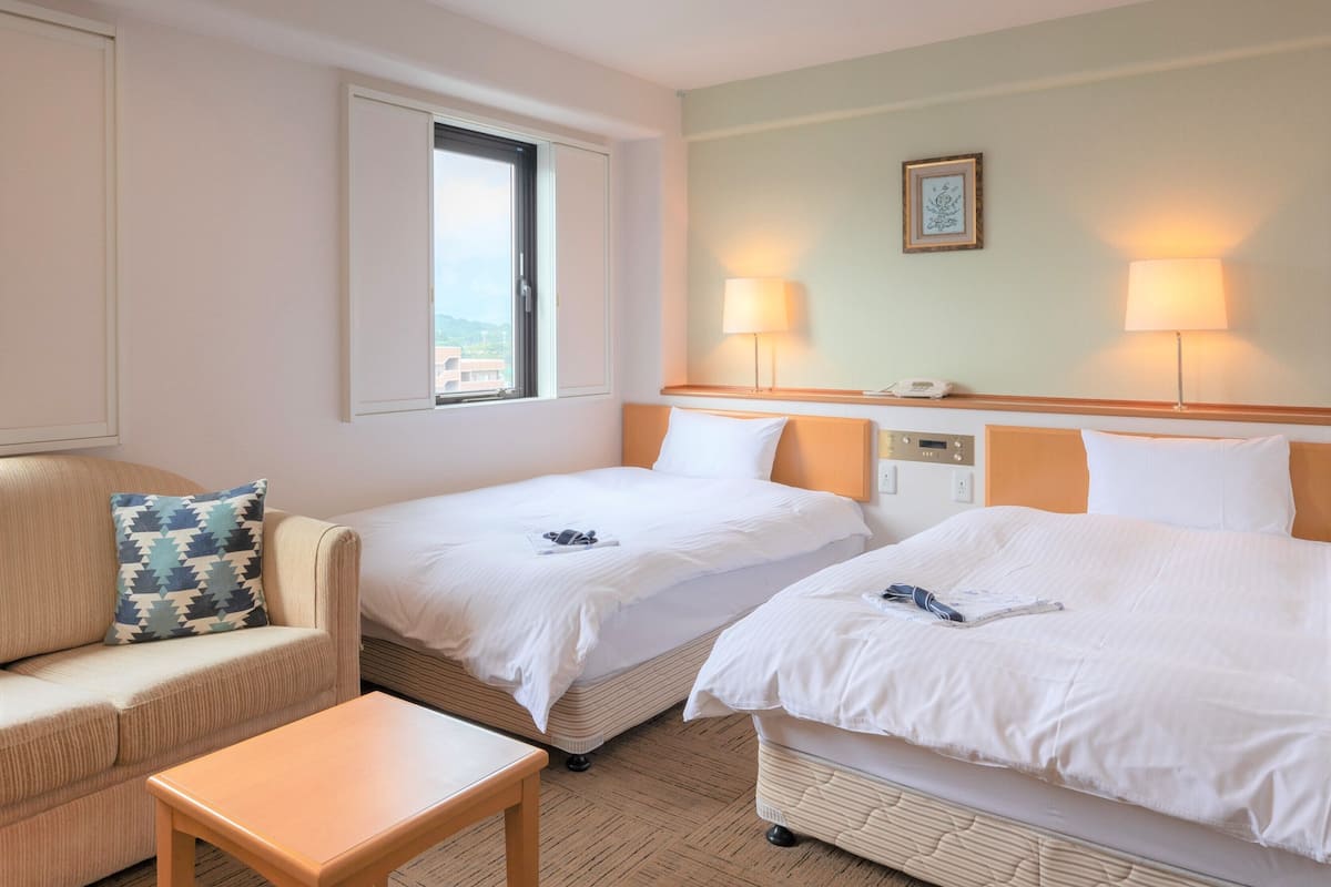 A hotel room with two beds, a sofa, a small table, and a window with a view.