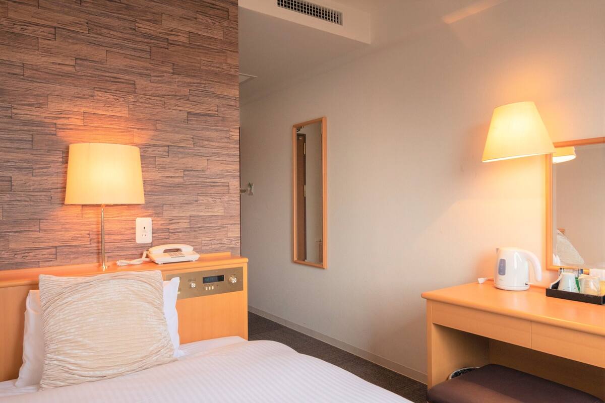 A hotel room with a bed, bedside table, lamp, and a desk with a chair.