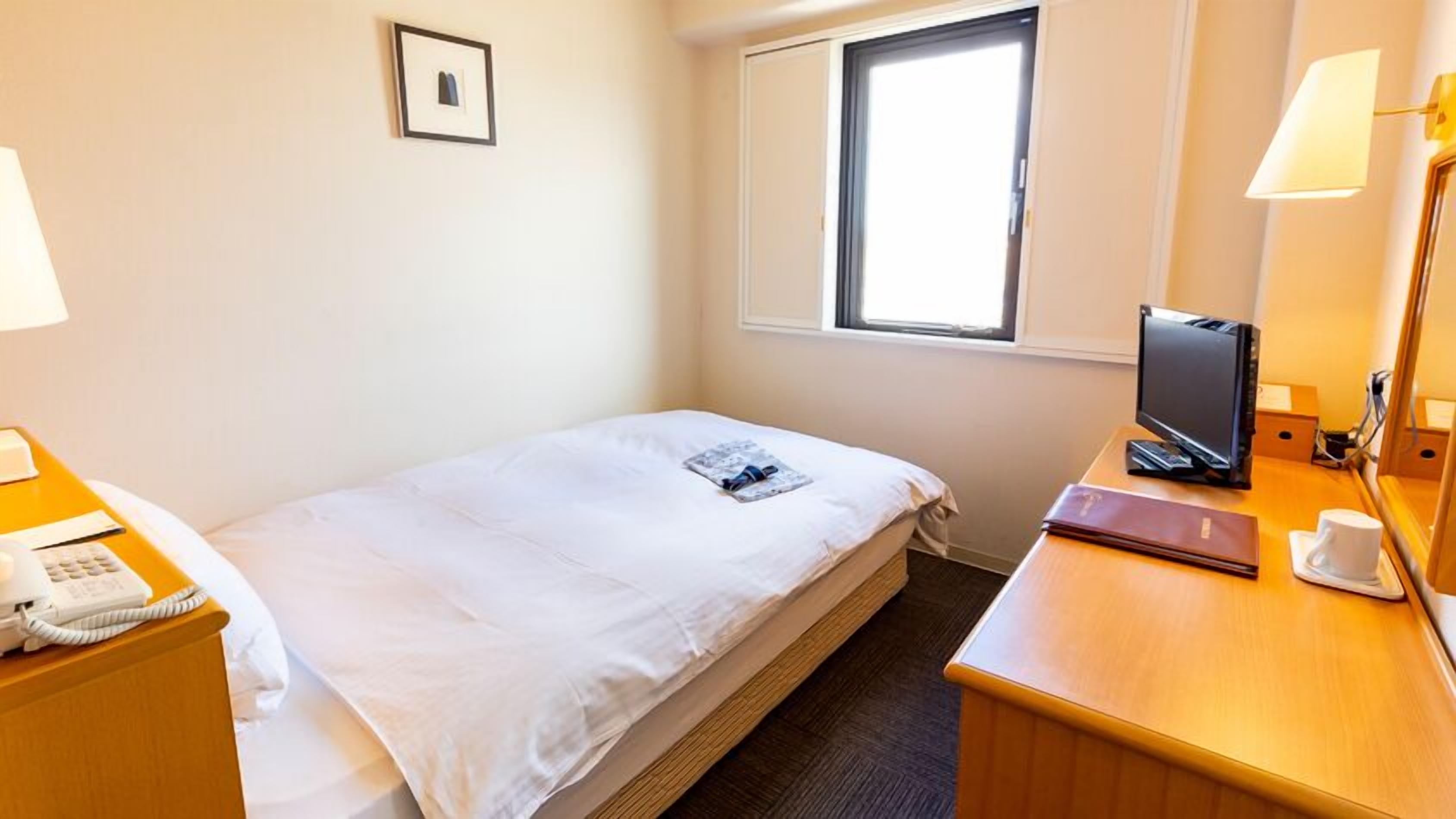 A hotel room with a bed, bedside table with a phone, a desk with a computer, a window, and a picture on the wall.
