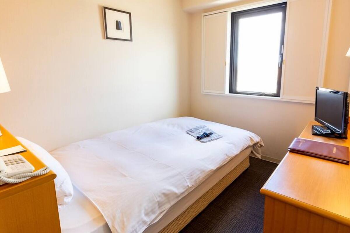 A hotel room with a bed, bedside table with a phone, a desk with a computer, a window, and a picture on the wall.