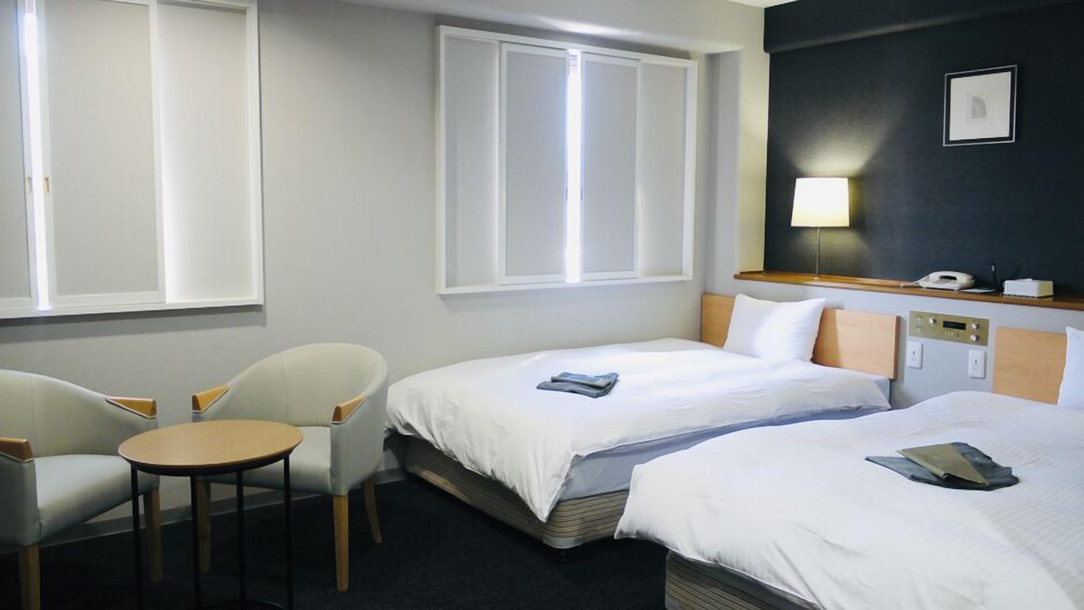 A hotel room with two beds, a small table, a chair, a bedside table with a lamp, and a wall-mounted phone.