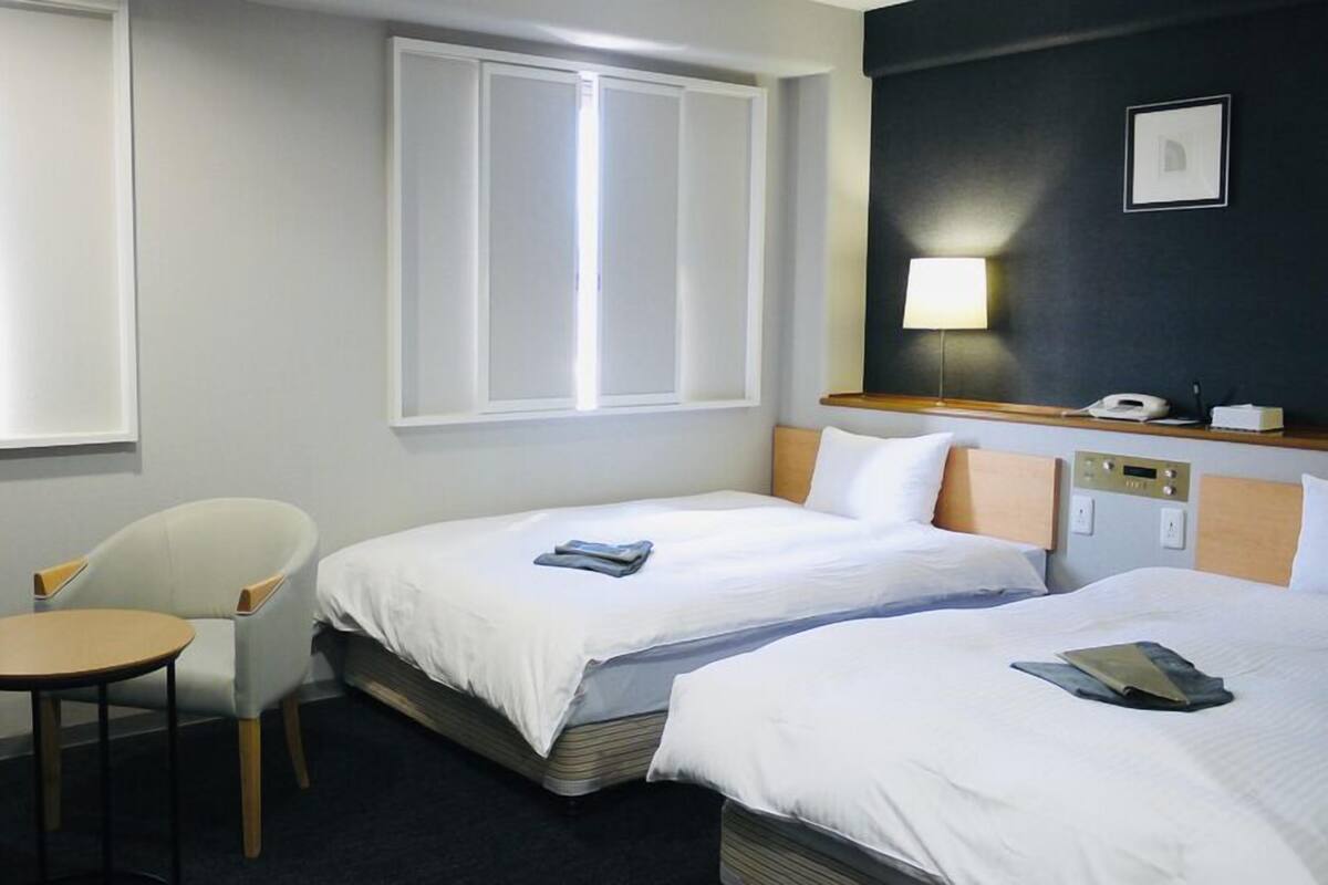 A hotel room with two beds, a small table, a chair, a bedside table with a lamp, and a wall-mounted phone.