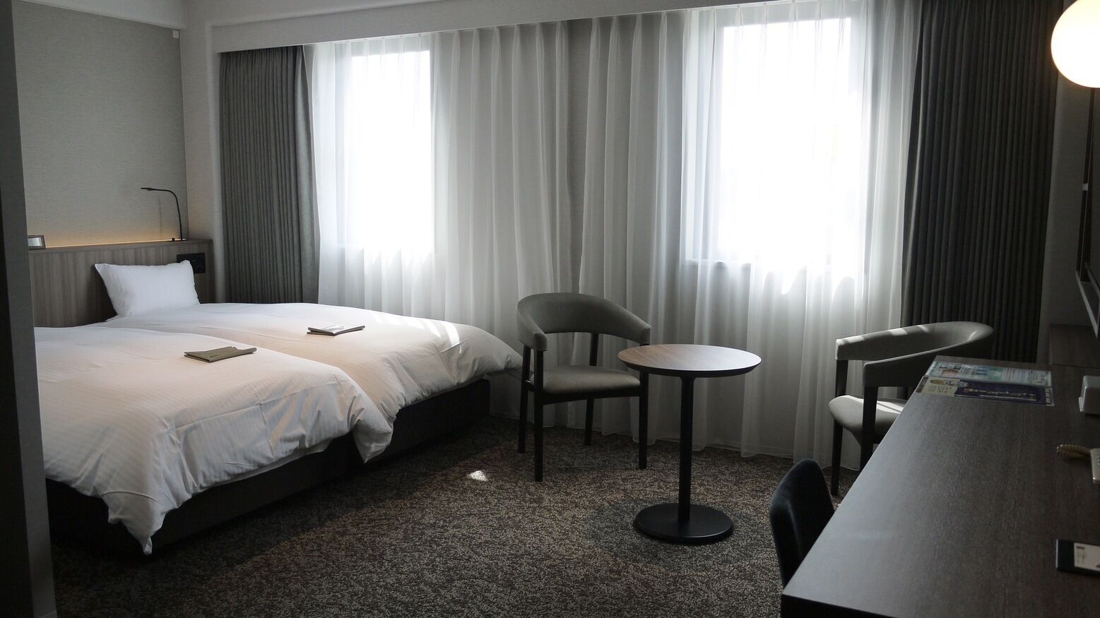 A hotel room with two beds, a small round table, two chairs, a desk, and a lamp.