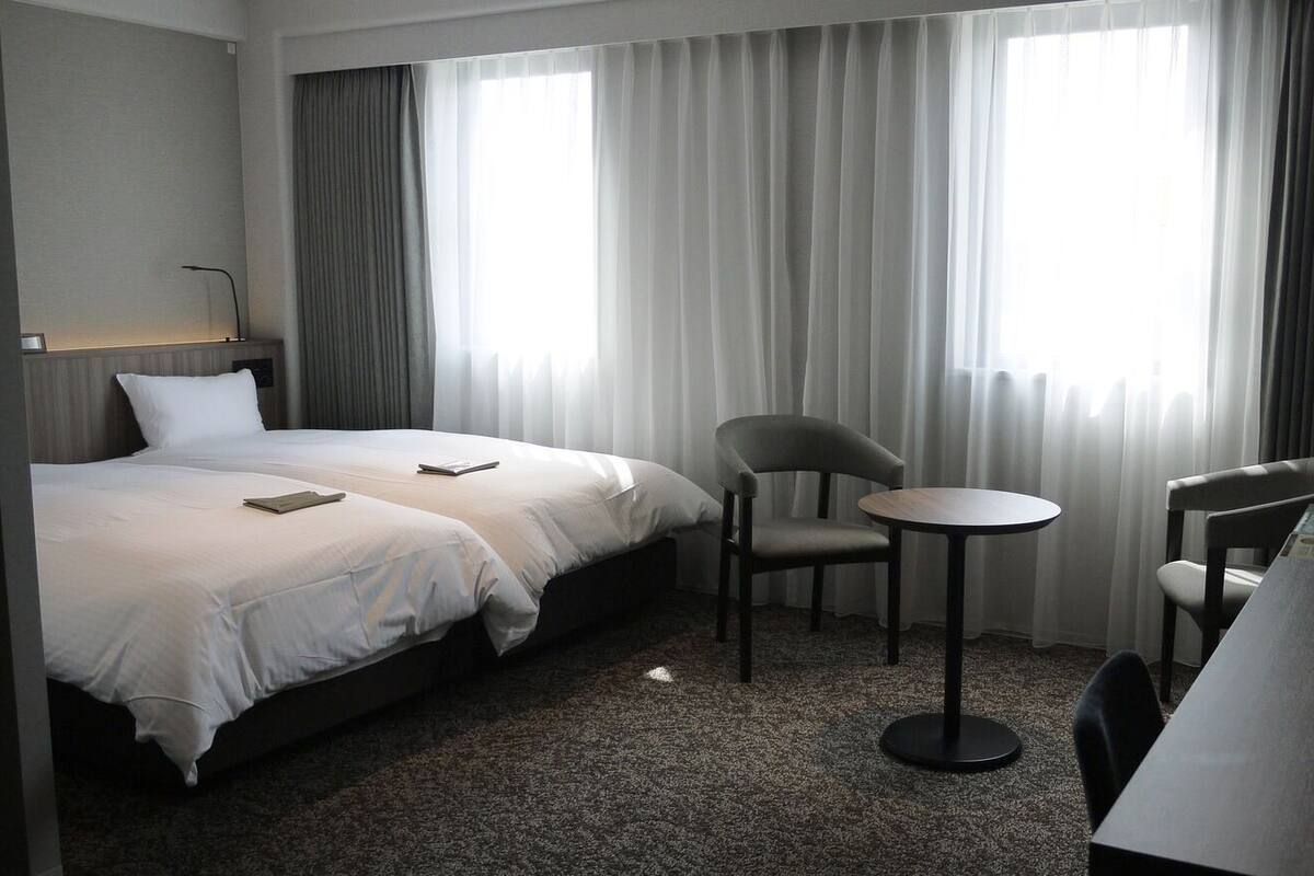 A hotel room with two beds, a small round table, two chairs, a desk, and a lamp.