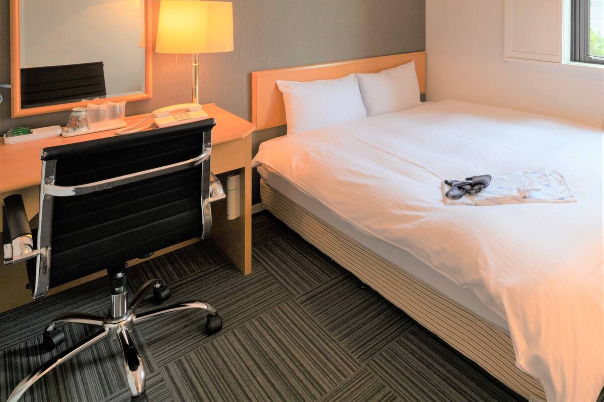 A hotel room with a bed, a desk with a lamp, a chair, and a mirror.