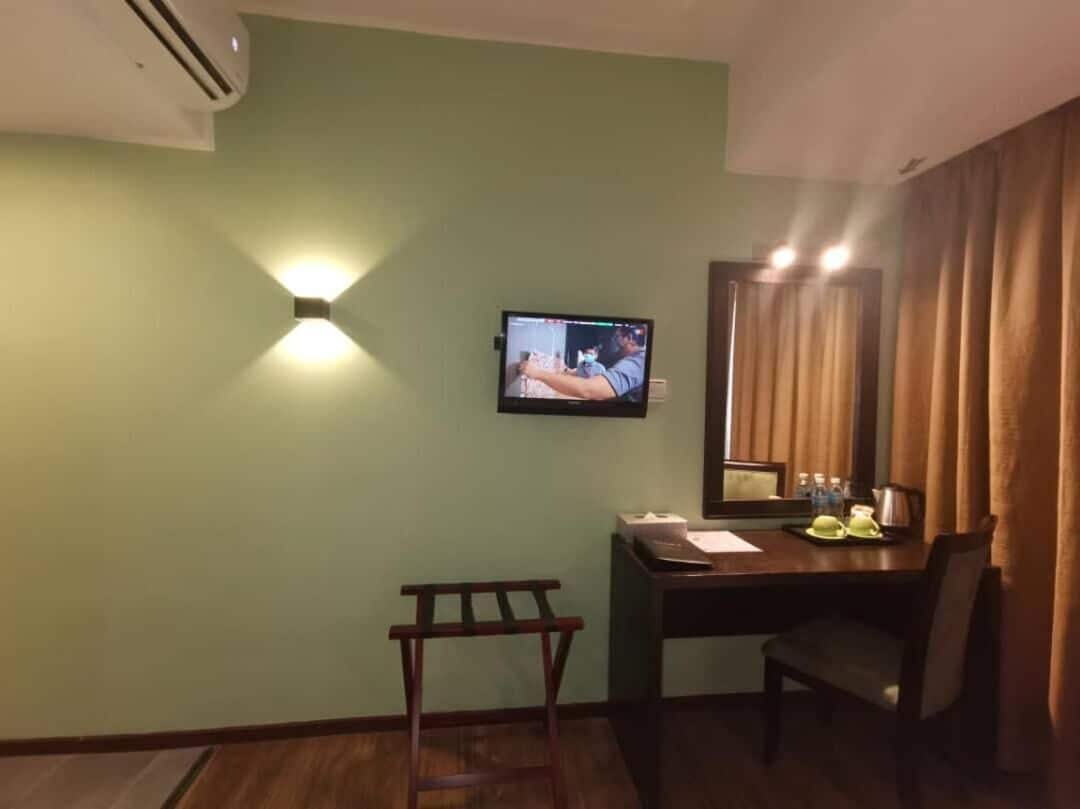 A hotel room with a television mounted on the wall, a desk with a chair, a small table, and a window with curtains.