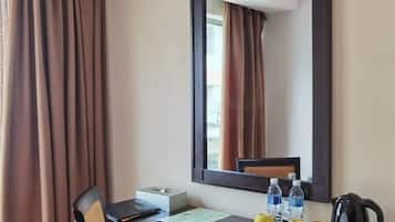 A hotel room with a desk, chair, air conditioning unit, mirror, and a window with curtains.