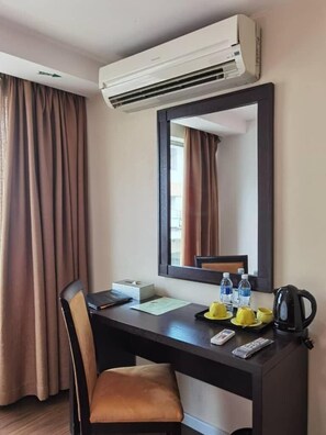 A hotel room with a desk, chair, air conditioning unit, mirror, and a window with curtains.