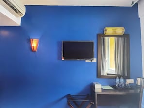 Une pièce avec un mur bleu, un téléviseur à écran plat fixé au mur, un luminaire mural et un miroir surmonté d’une fenêtre.