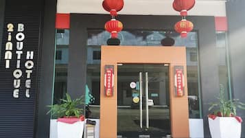 The entrance of a hotel with a black facade, glass doors, and red lanterns above. There are two large potted plants at the entrance.