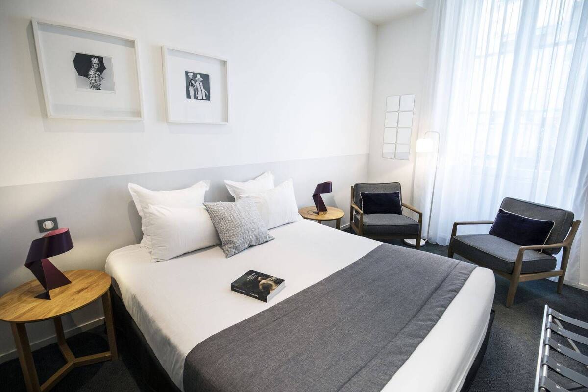 Une chambre d’hôtel avec un grand lit, deux fauteuils, une petite table avec une lampe et deux tableaux encadrés au mur.