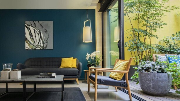 Un salón moderno con pared azul oscuro, un sofá gris con cojines amarillos, una silla de madera, una mesa de centro y una planta grande en maceta.