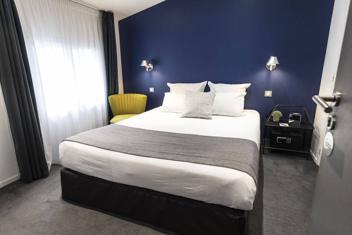 Une chambre d’hôtel avec un grand lit, une table de chevet avec une lampe, une chaise et une petite table avec une plante.
