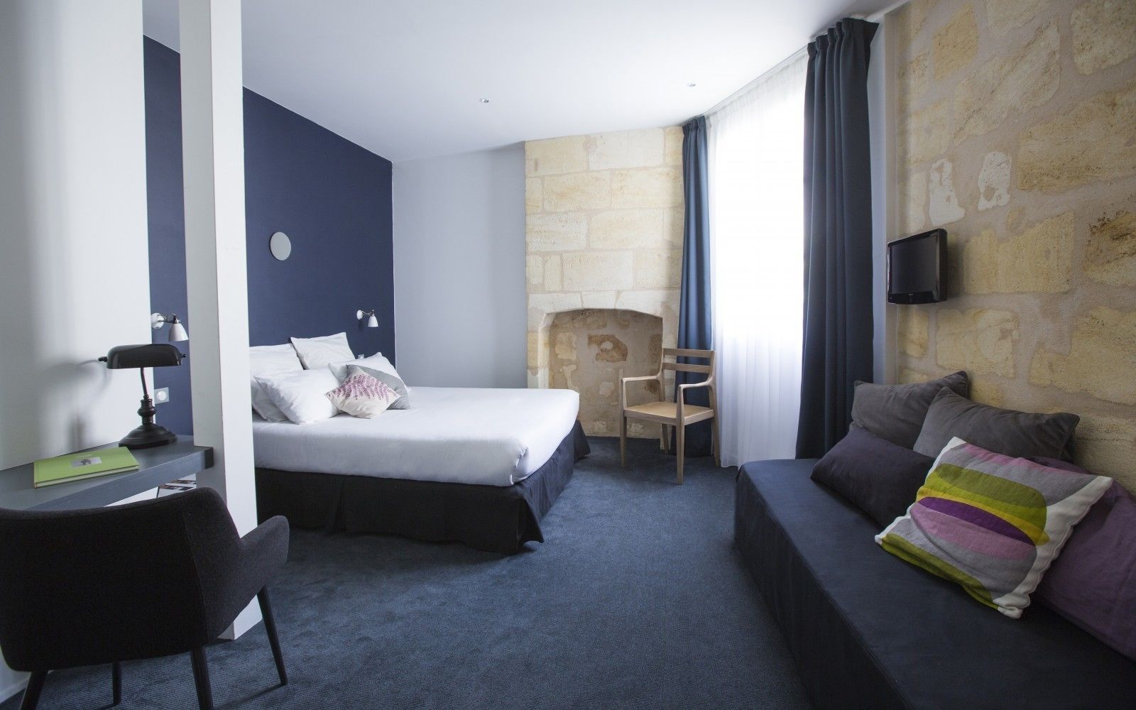 A hotel room with a bed, a desk with a lamp, a chair, a sofa, and a stone wall.