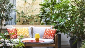 Un rincón al aire libre acogedor con un banco de madera y cojines, rodeado de abundante vegetación y plantas en macetas.