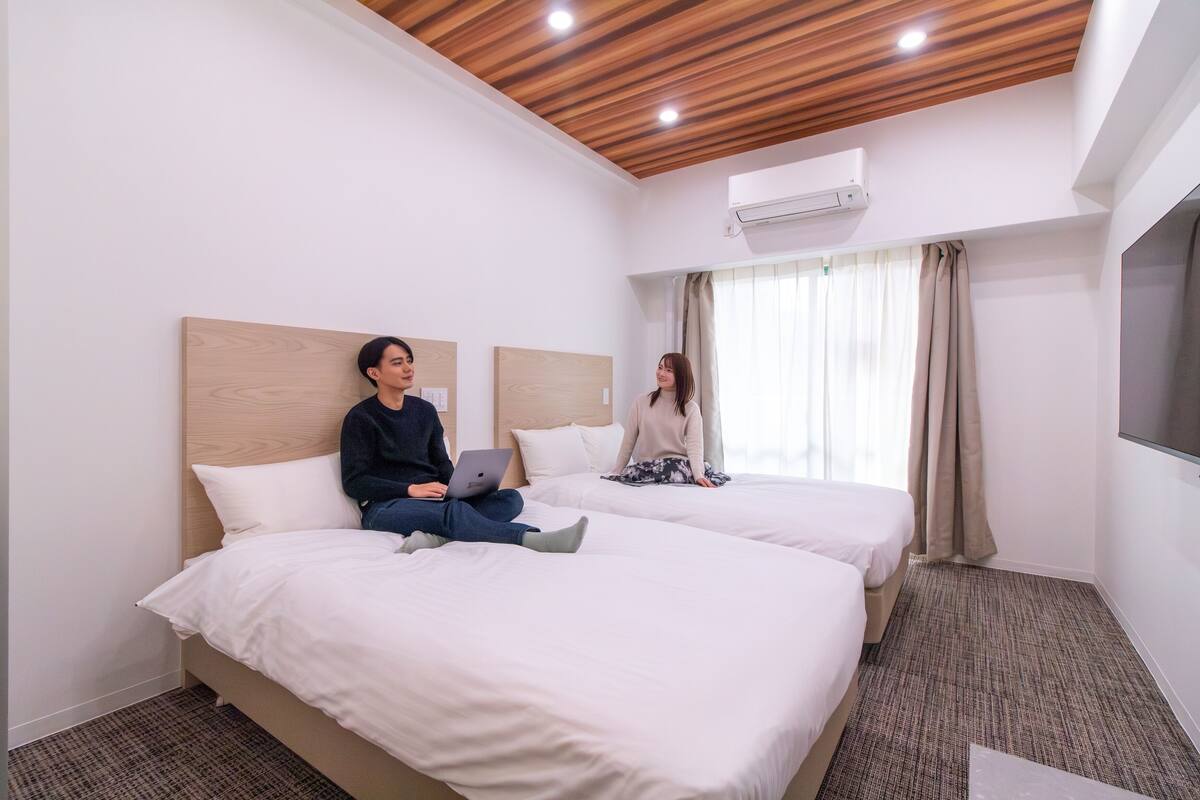 A hotel room with two beds, a wooden ceiling, an air conditioning unit, and a large window with curtains.