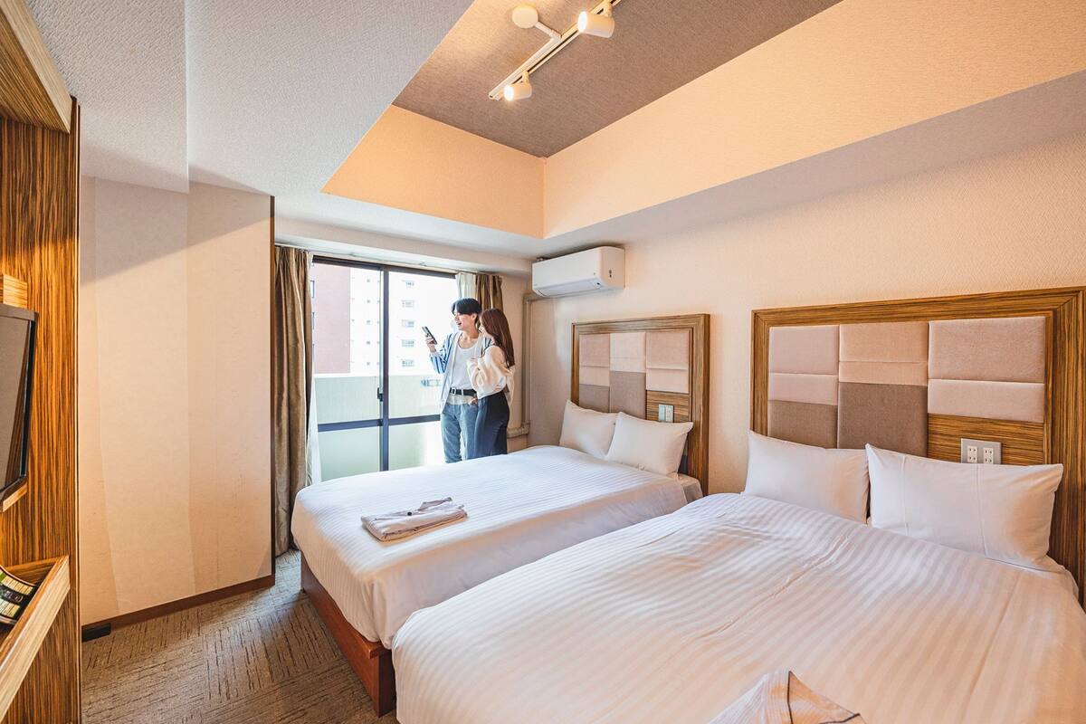 A hotel room with two beds, a wooden headboard, a large window, and an air conditioning unit.