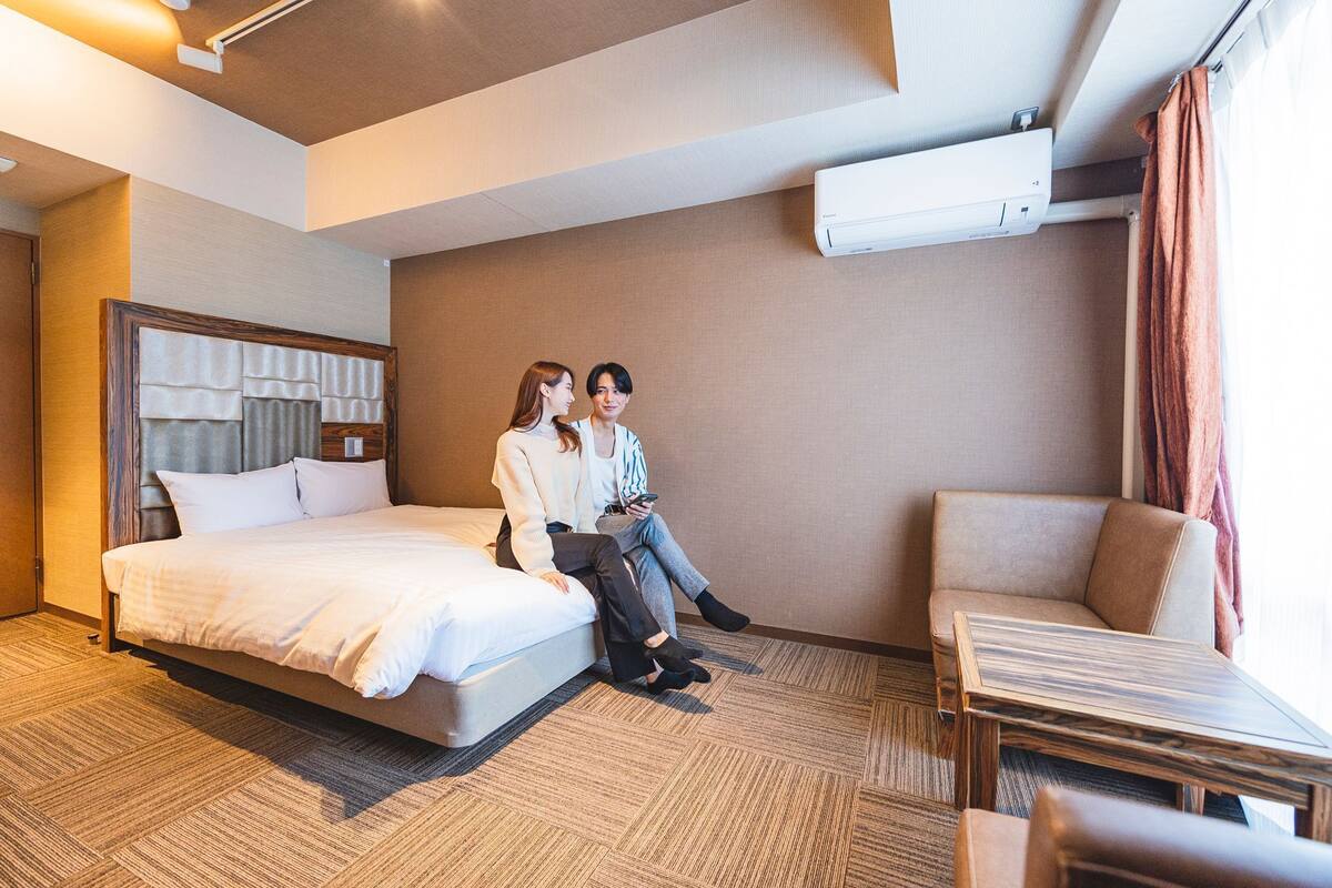 A hotel room with a bed, two people sitting on the bed, a chair, and a table.