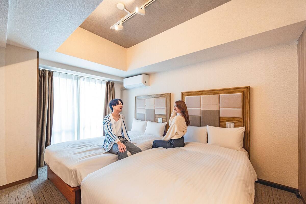 A hotel room with two beds, a large window with curtains, a ceiling light fixture, and a wall-mounted air conditioning unit.