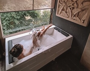 Una persona en una bañera con una ventana arriba, rodeada de un muro de azulejos con un diseño labrado.
