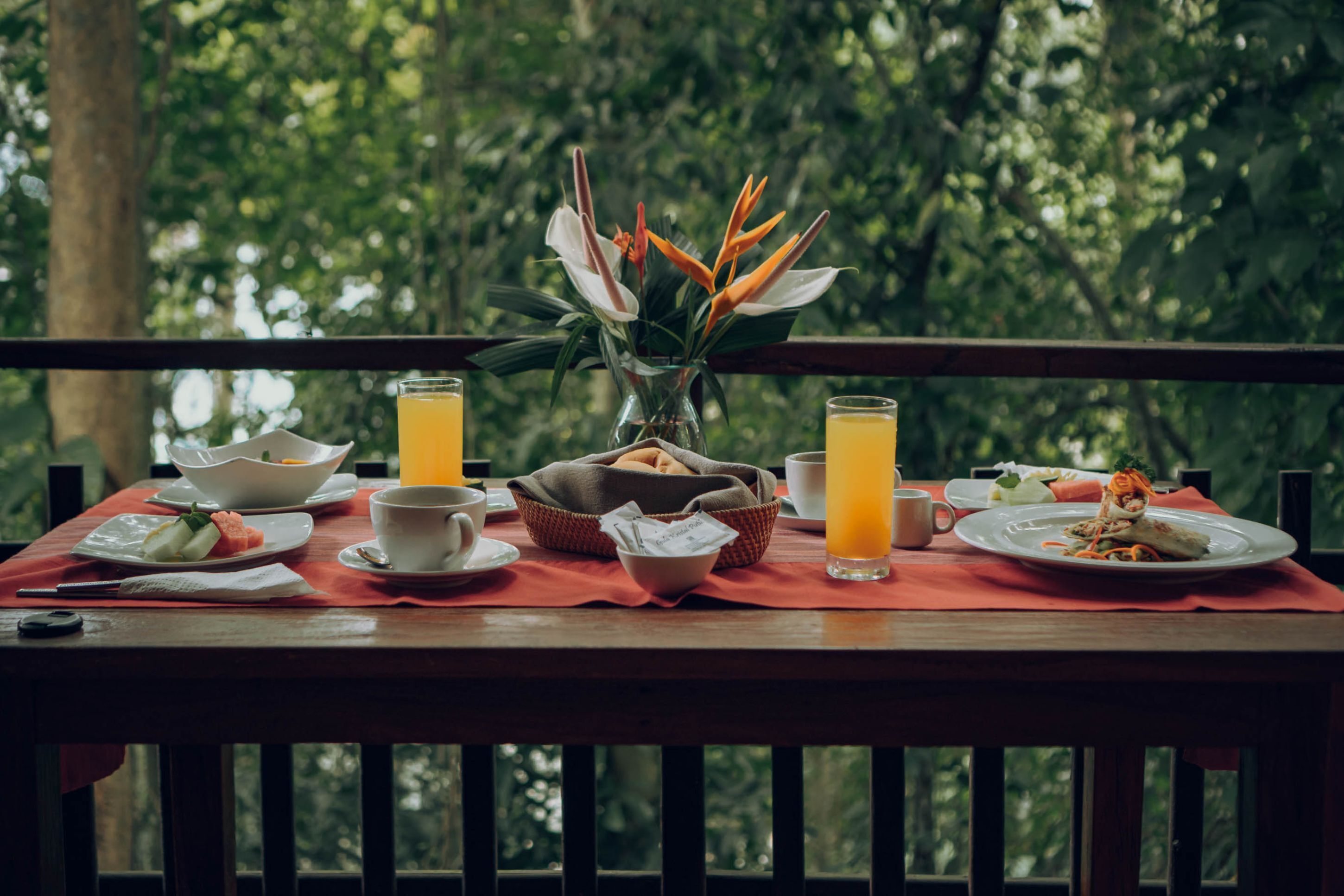 A dining table set with breakfast items, including a bowl, cups, and a vase with flowers, on a balcony with a forest view.