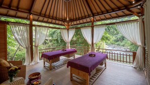 A wooden deck with massage tables, wicker furniture, and a view of a river. - Adiwana Svarga Loka (Ubud)