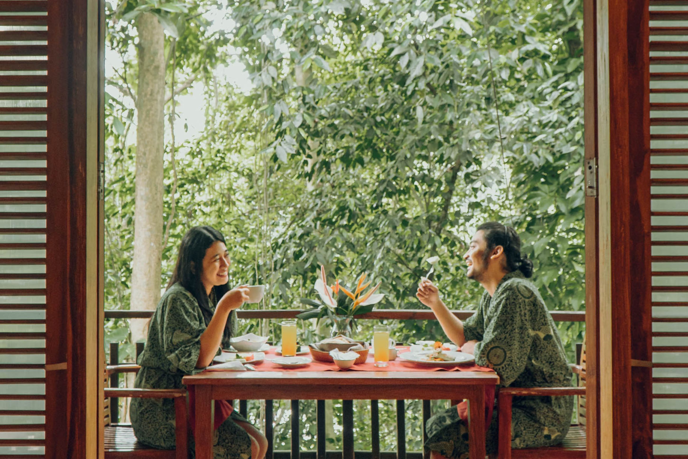 A wooden table with a red tablecloth, surrounded by people in patterned clothing, with a lush green background visible through a wooden frame.
