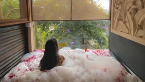 A person sitting in a bathtub filled with bubbles and rose petals, with a view of trees and flowers through a window. - Adiwana Svarga Loka (Ubud)