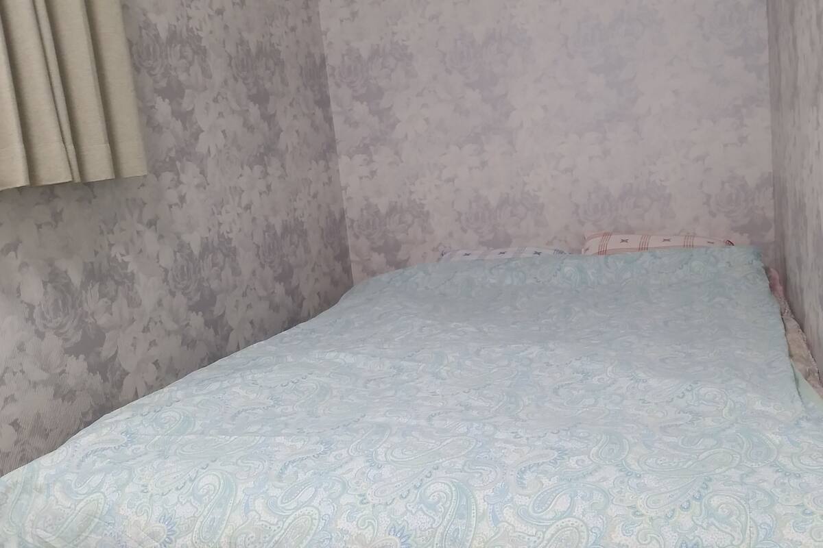 A small, narrow room with a single bed. The walls are plain and unadorned.