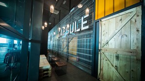 Un interior moderno con una puerta grande de madera con la etiqueta “CAPSULE”, un separador de vidrio y un área de descanso con mesas y sillas.