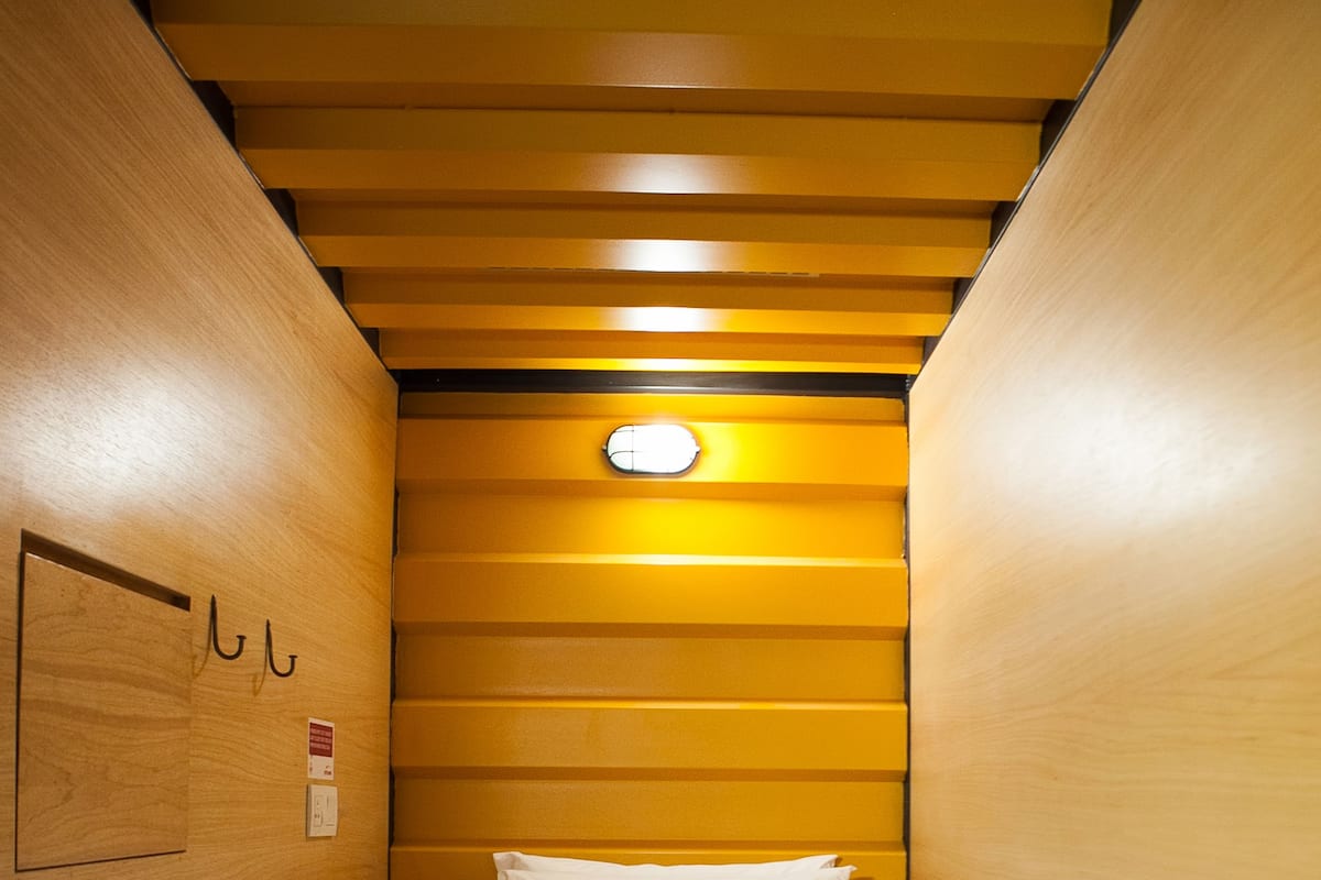 A narrow, wooden-paneled room with a single bed, a small wall-mounted light, and a simple electrical outlet.