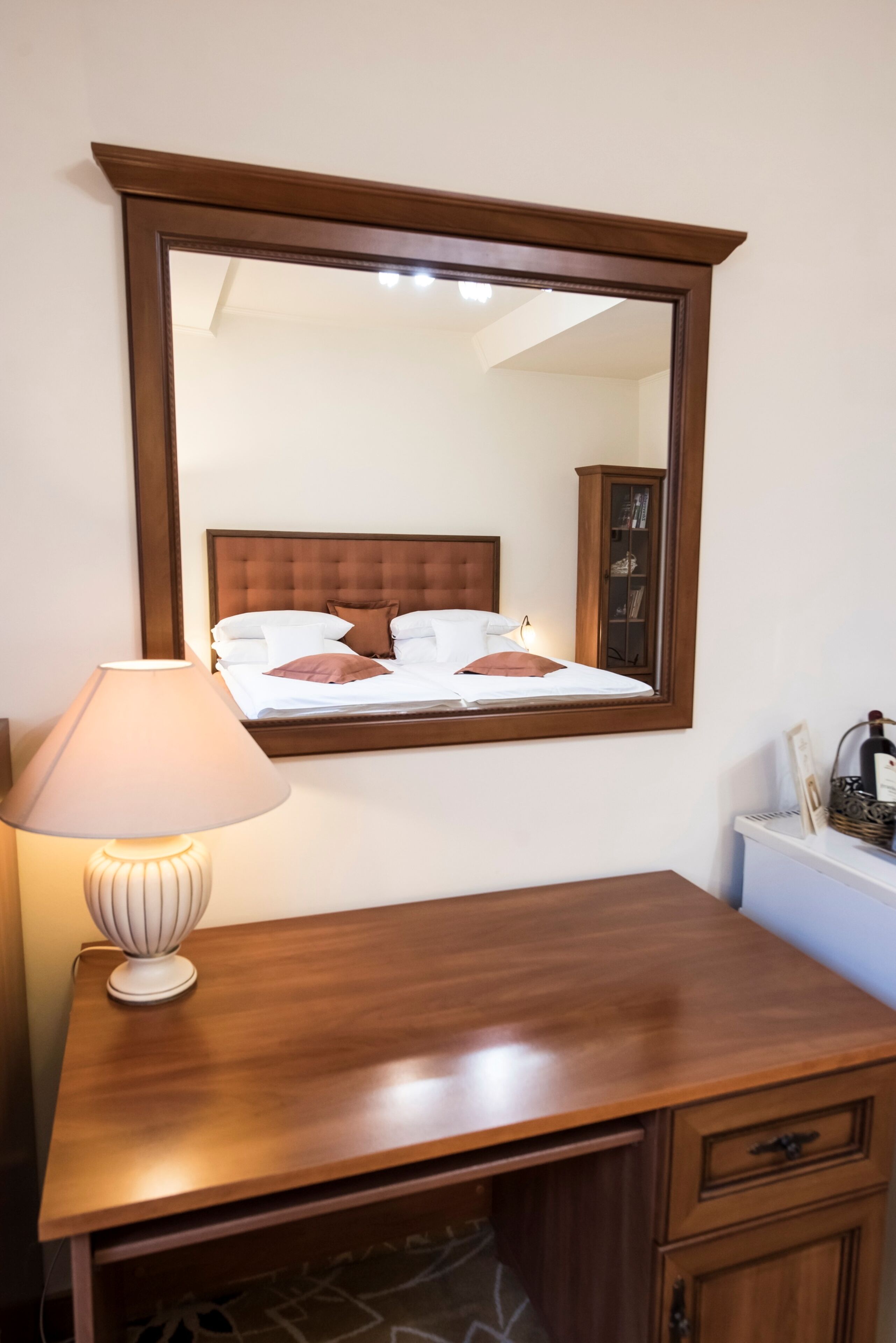 A wooden desk with a lamp, a mirror above it, and a bed with a headboard in the reflection.