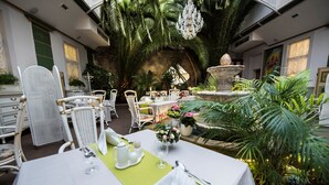 Un comedor con mesas dispuestas para una comida, rodeado de abundante follaje y una fuente decorativa.