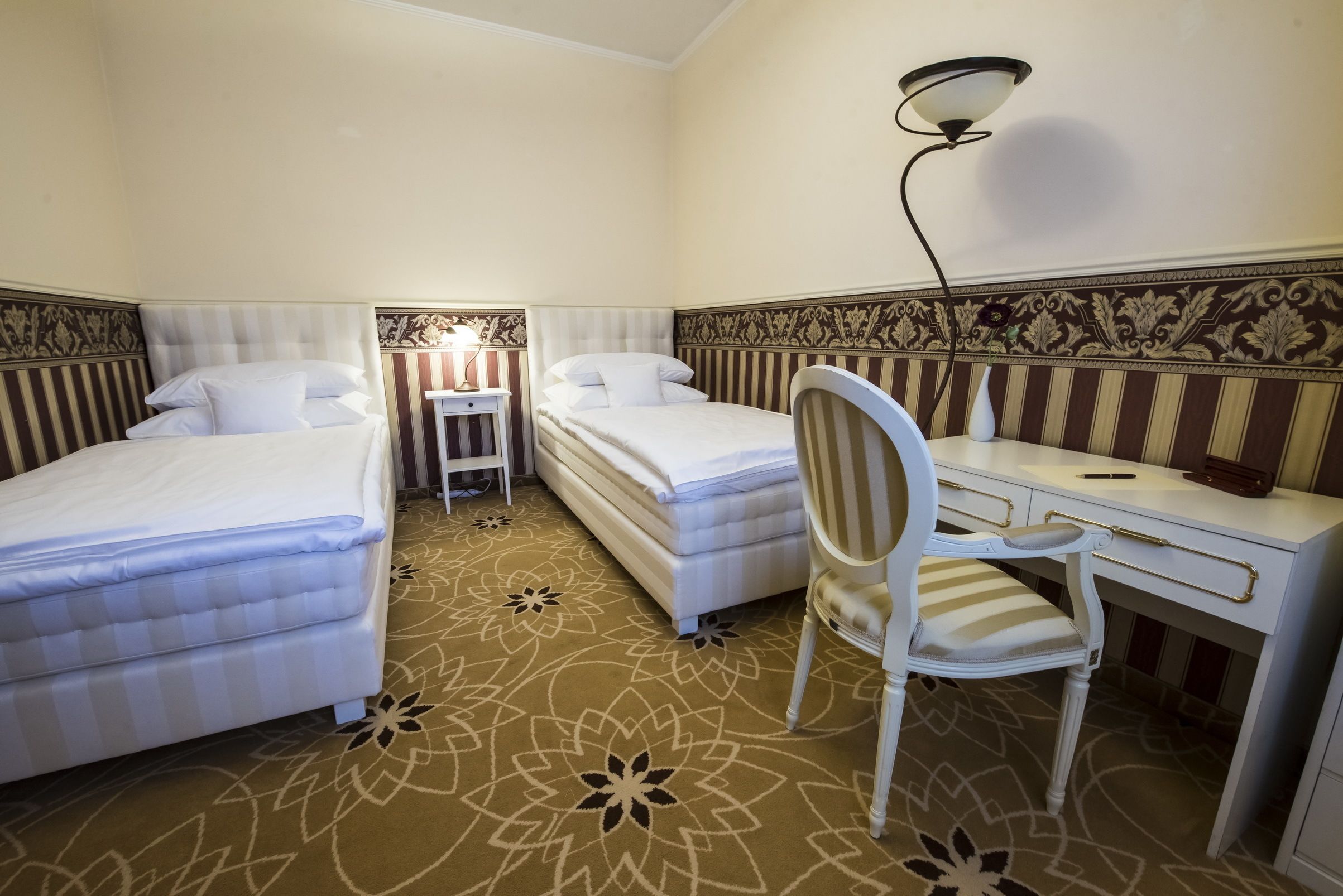 Une chambre d’hôtel avec deux lits, un bureau, une chaise, une lampe et une moquette à motifs.