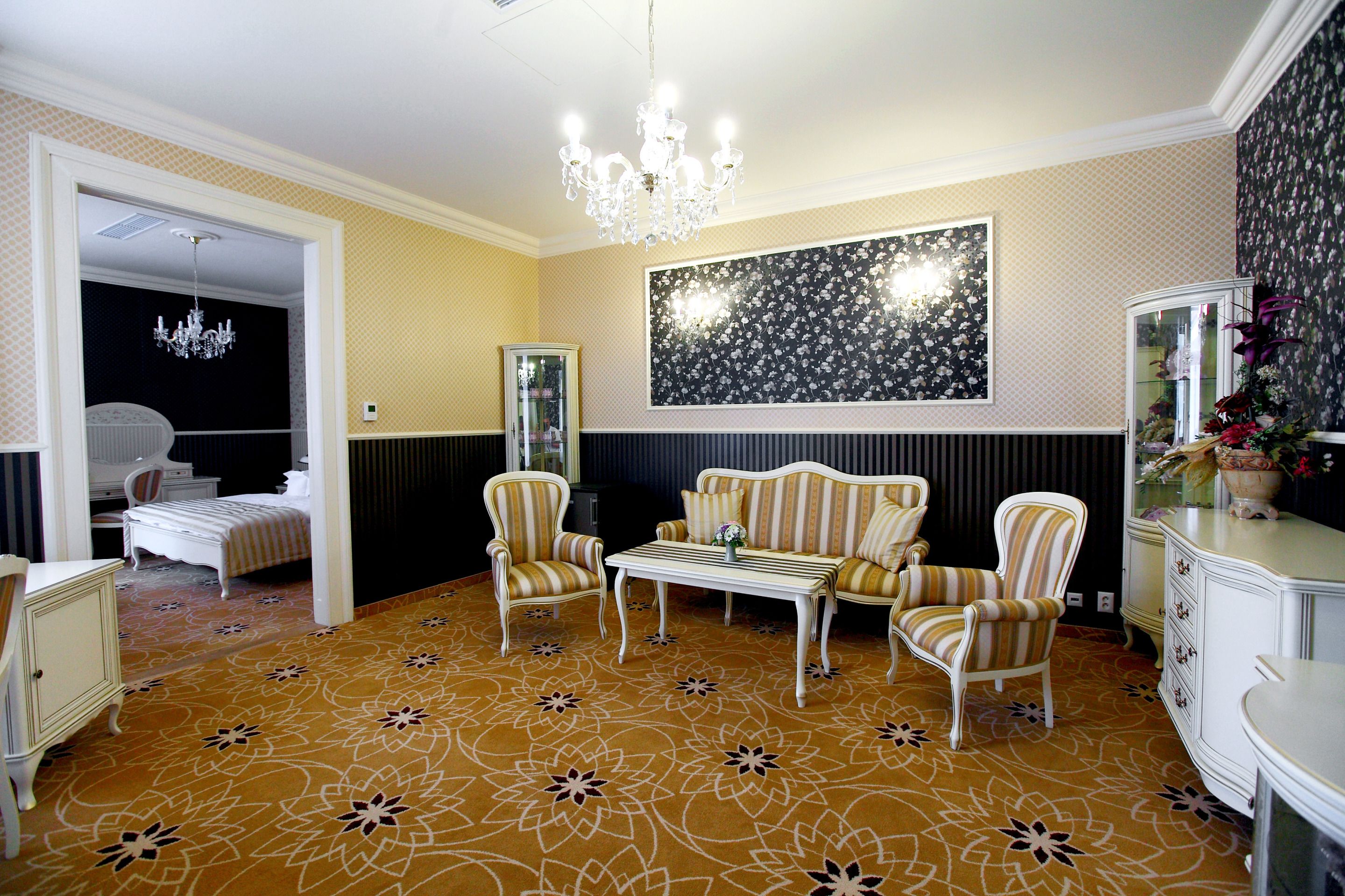 A room with a patterned carpet, a chandelier, striped armchairs, a white table, and a large floral artwork on the wall.