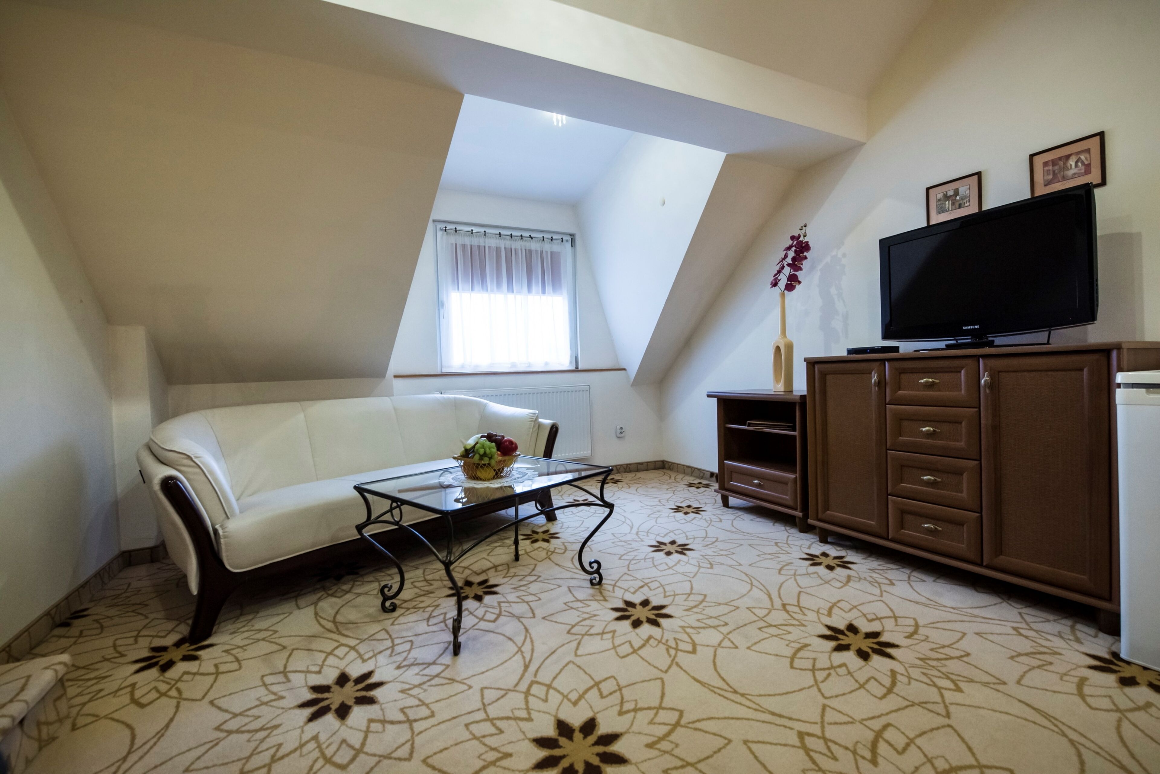 A living room with a white sofa, a glass coffee table with fruit, a TV on a wooden cabinet, and a patterned carpet.