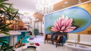 Un área de piscina interior lujosa, con un gran mural de loto, un jacuzzi y asientos junto a la piscina.