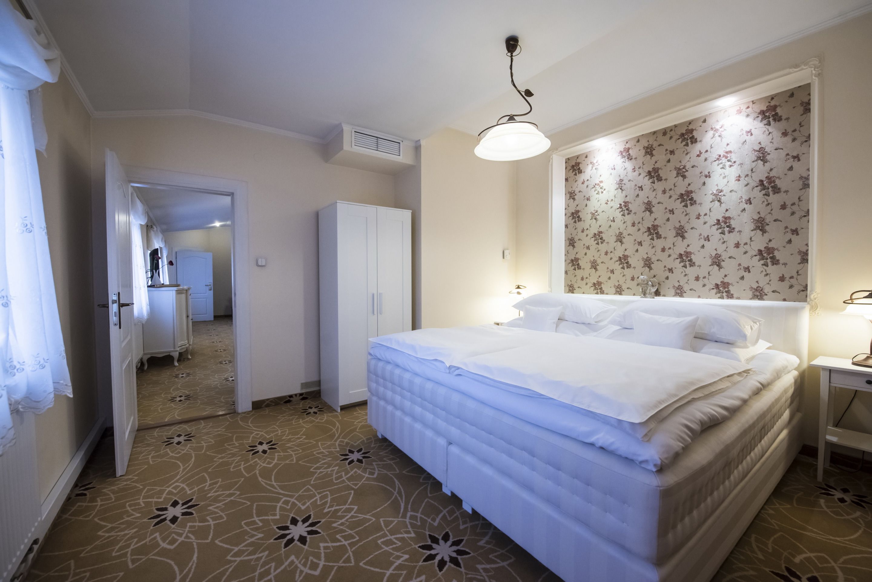 A hotel room with a bed, a wardrobe, a mirror, and a floral wallpaper.