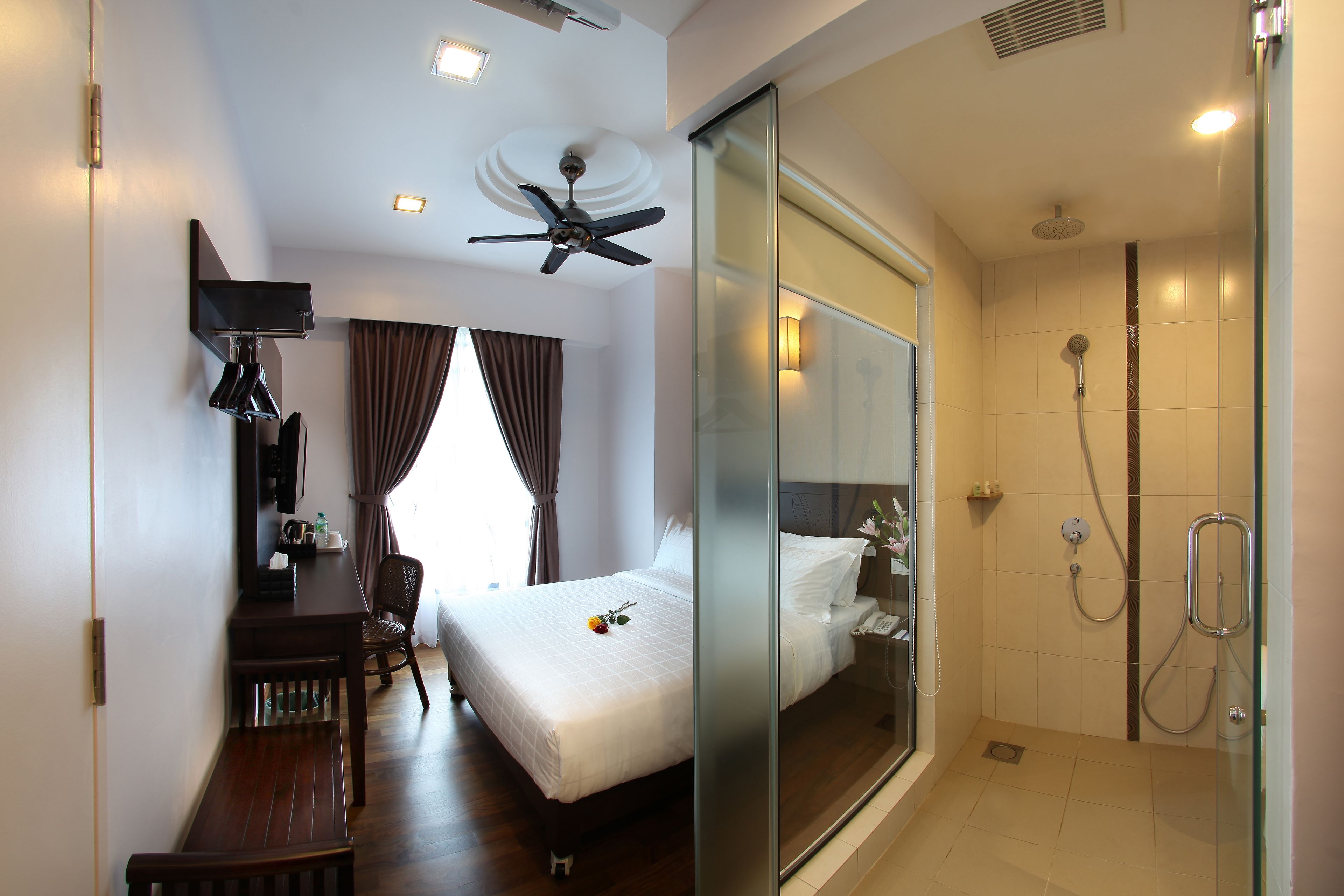 A hotel room with a bed, a desk, a chair, a ceiling fan, a shower area, and a glass partition.