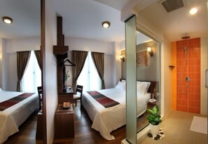 Une chambre d’hôtel avec deux lits, un bureau, une chaise, une petite table, une plante en pot et un espace douche avec un mur recouvert de carreaux orange.