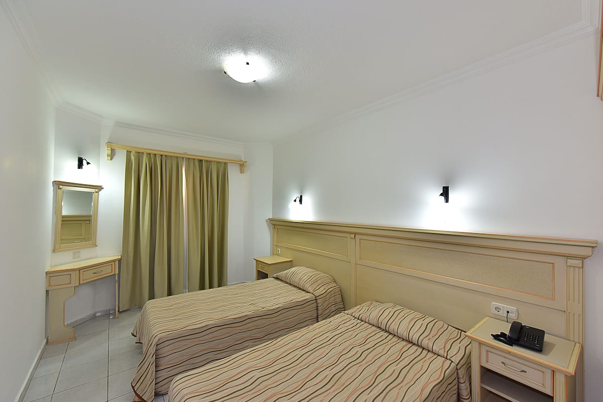 A hotel room with two beds, a bedside table with a phone, a nightstand, a mirror, and a window with curtains.